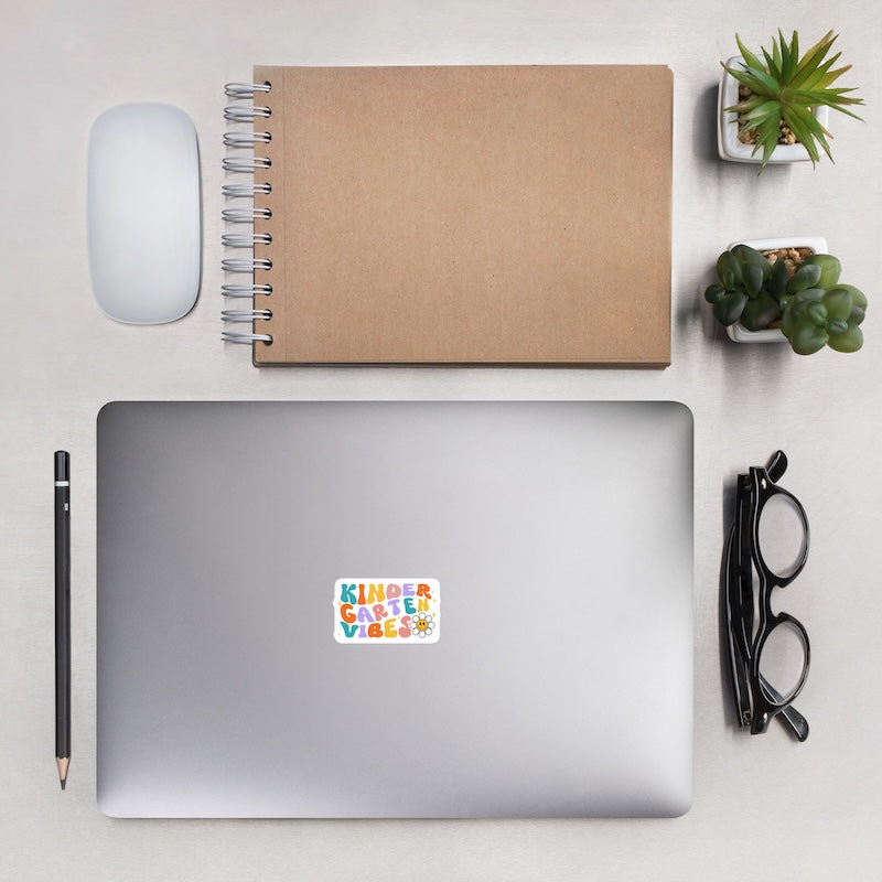 Laptop with a colorful sticker, notebook, mouse, pen, glasses, and small plants on a light surface.