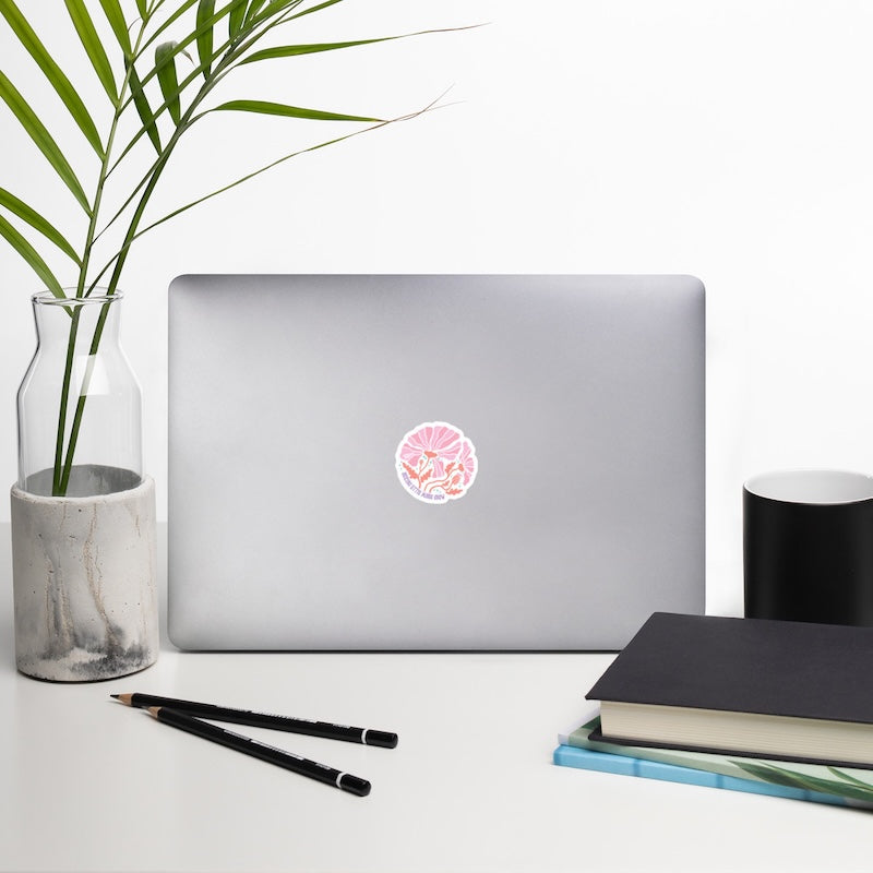 Laptop with a decorative sticker on a desk with a plant, pencils, and books.