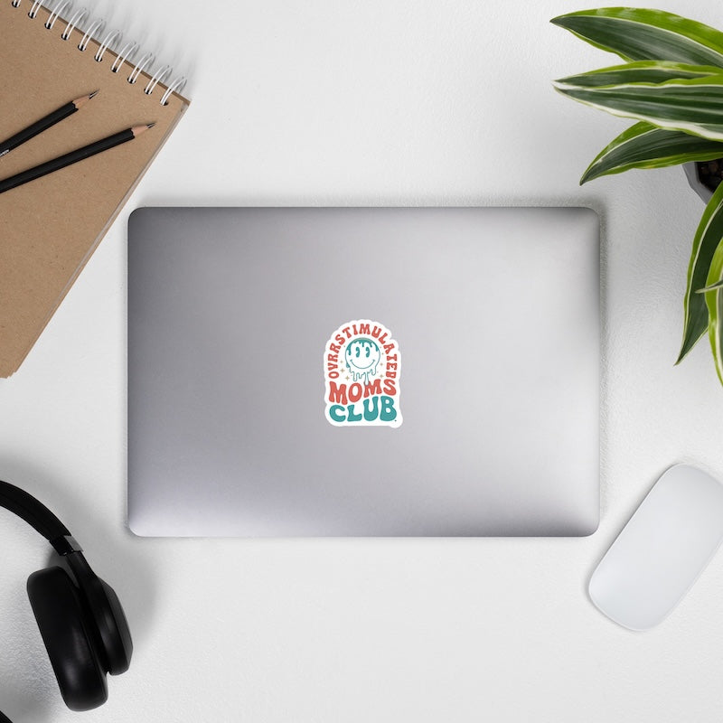 Laptop with a 'Mom's Club' sticker on a desk with a notebook, pencils, headphones, and a plant.