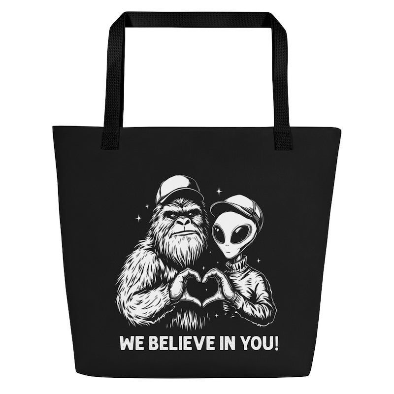 Black tote bag with a design of a bigfoot and alien making a heart shape with their hands and text 'WE BELIEVE IN YOU!' on a white background.