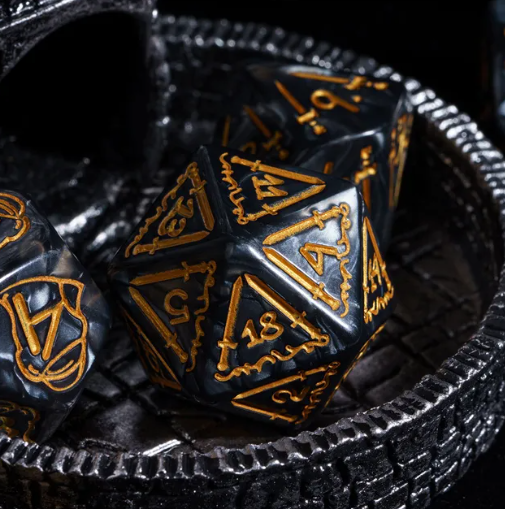Black polyhedral dice with gold numbering and symbols on a textured black surface