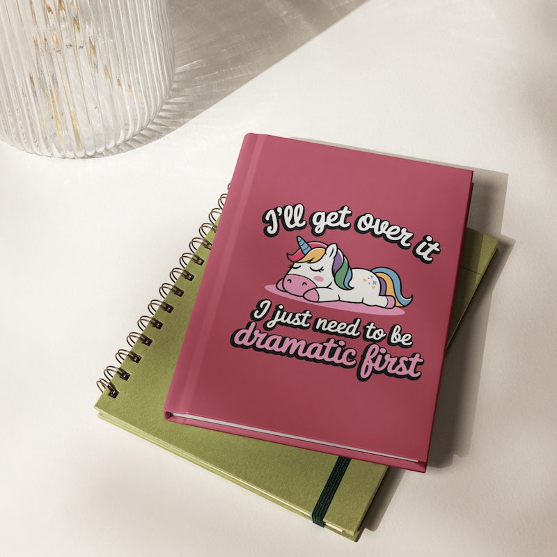 Pink notebook with unicorn illustration and humorous text on a white surface