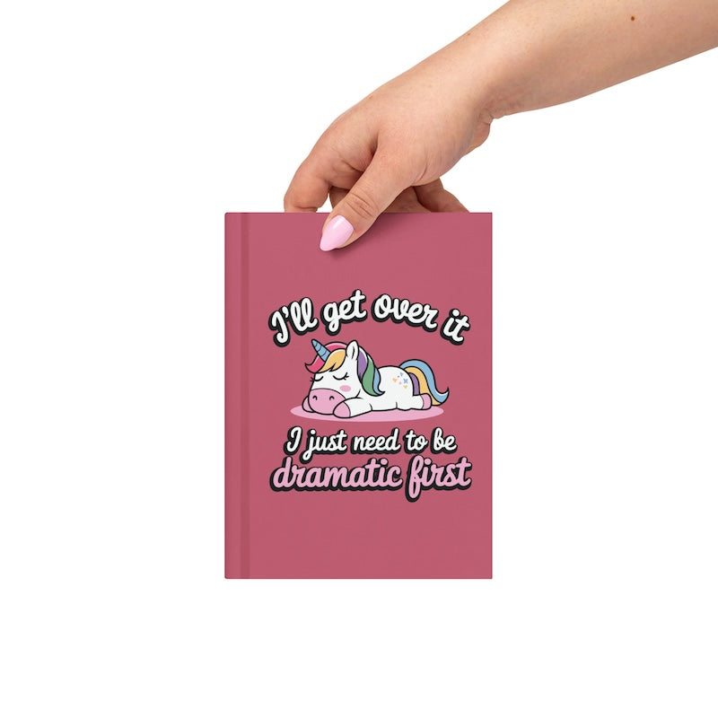 Pink notebook with unicorn graphic and text, held by a hand on a white background