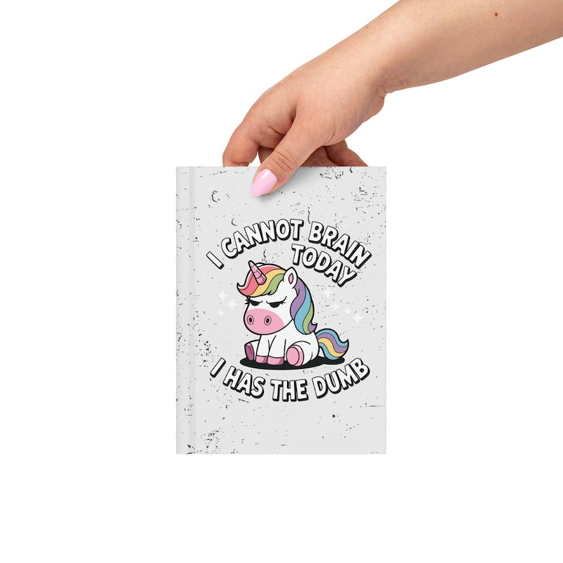 Notebook with a unicorn illustration and humorous text held by a hand on a white background