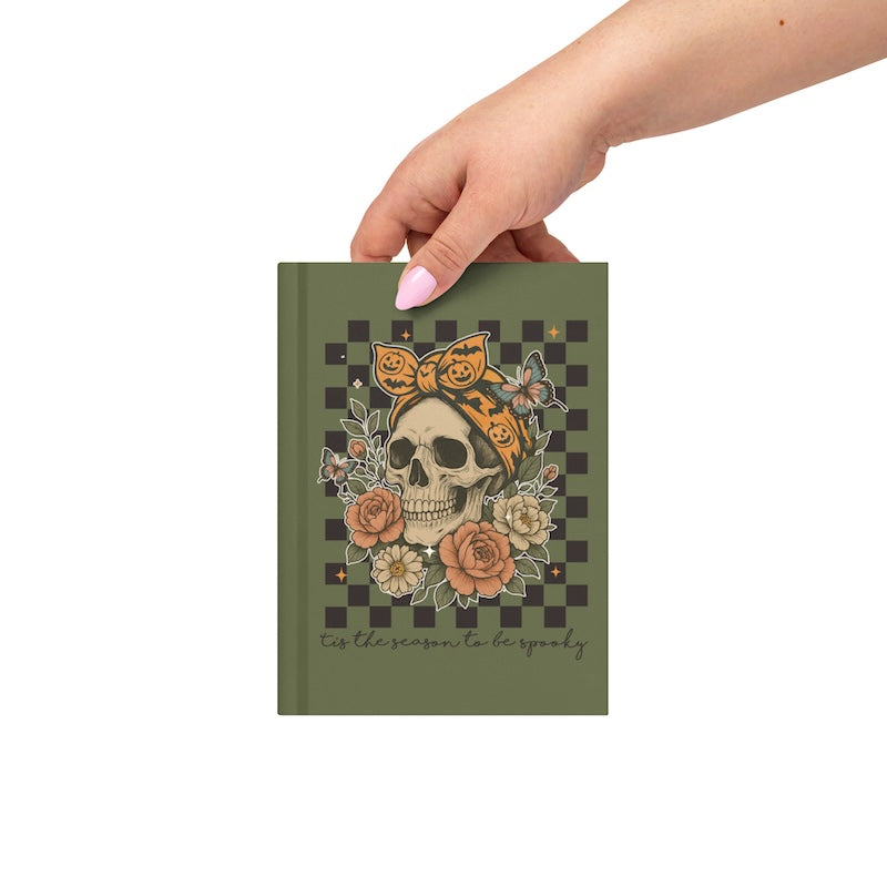 Hand holding a notebook with a skull and floral design on a checkered background