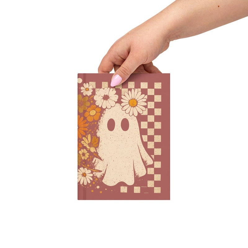 Hand holding a notebook with a ghost and floral design on a checkered background