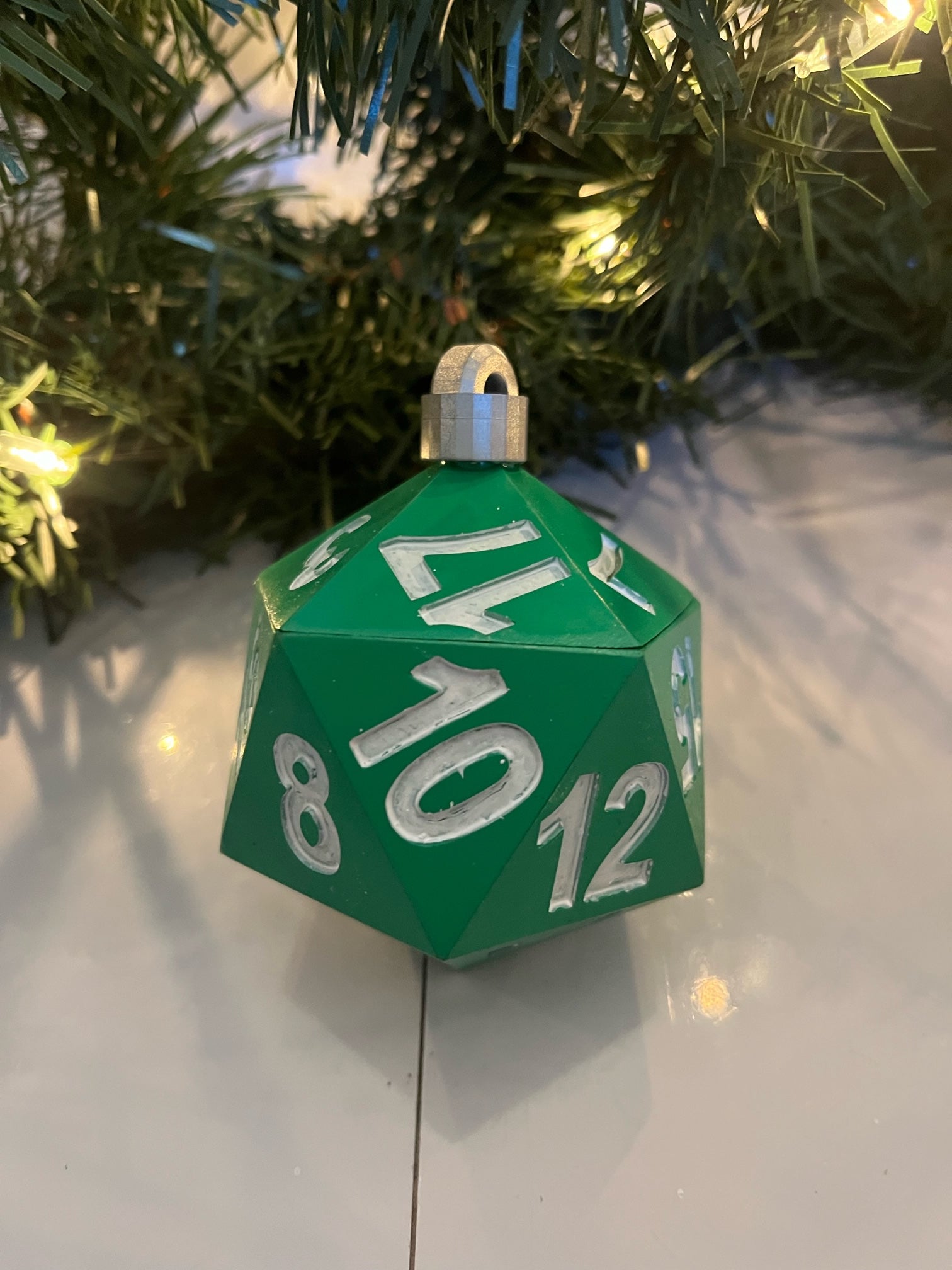 Green polyhedral die ornament with numbers on a reflective surface with a Christmas tree in the background.