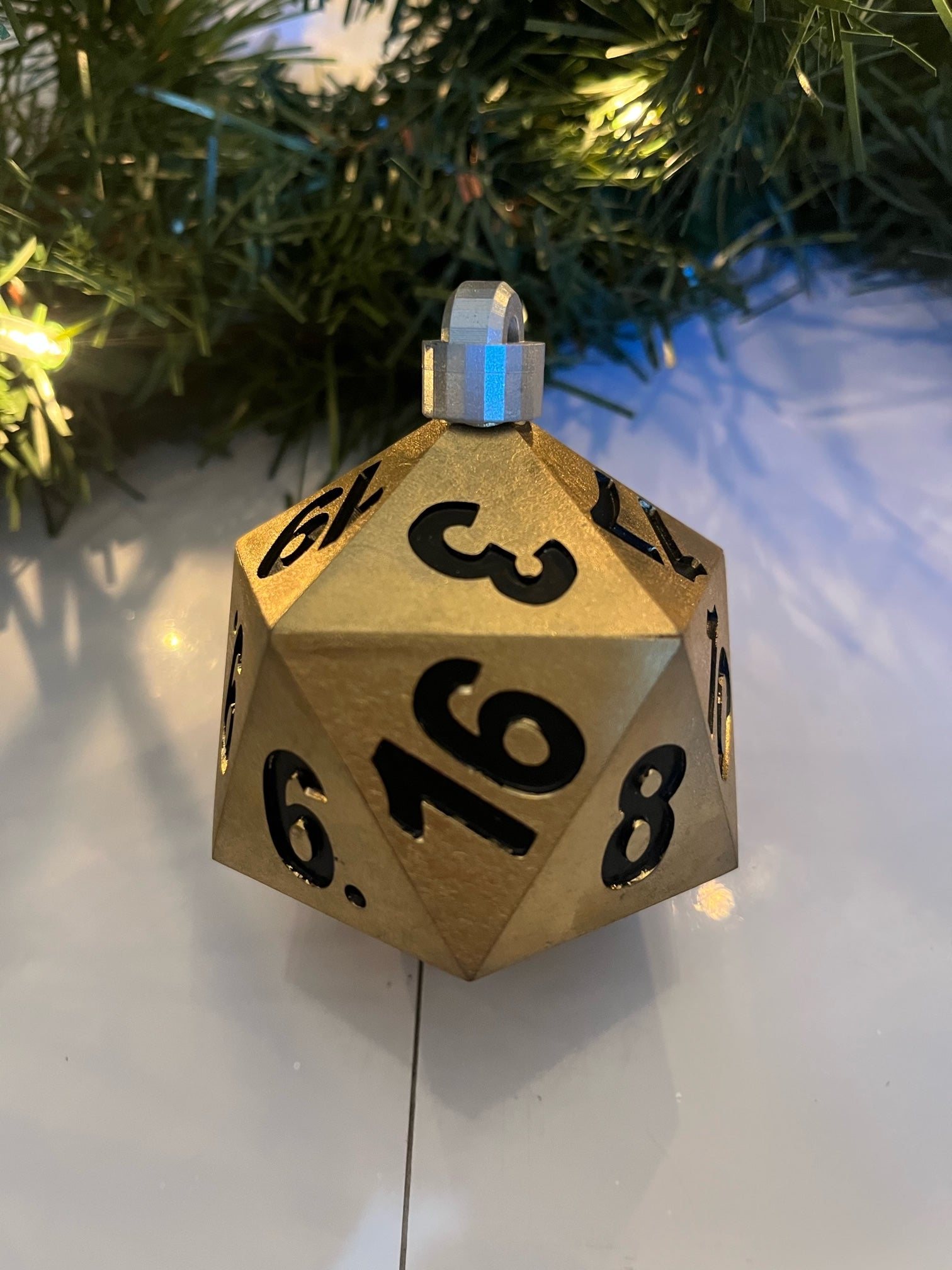 Gold D20 dice ornament with black numbers on a white surface with Christmas tree in background