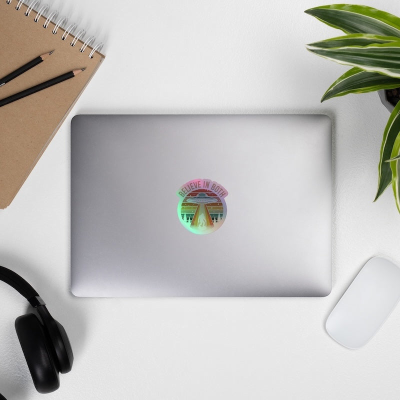 Silver laptop with a colorful sticker on a white surface with a notebook, pencils, headphones, and a plant.