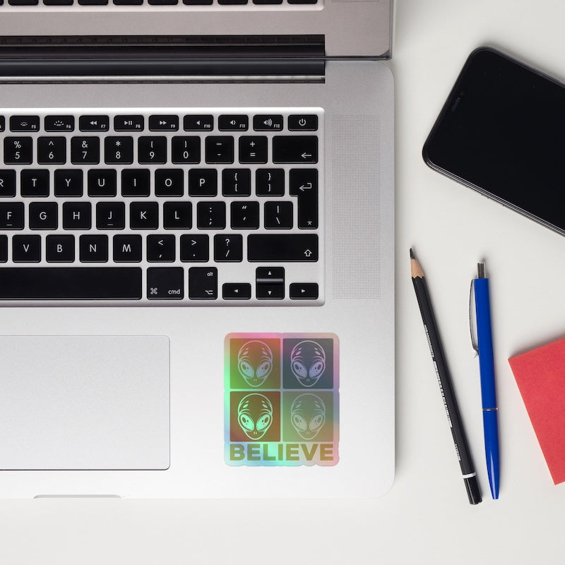 Laptop with a sticker featuring aliens and the word 'BELIEVE', next to a smartphone, pencil, and pen on a white surface.