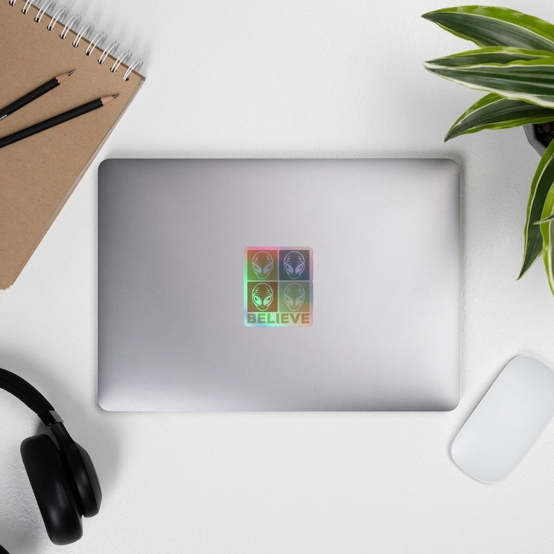 Silver laptop with a colorful sticker on a white surface with a plant and office supplies.