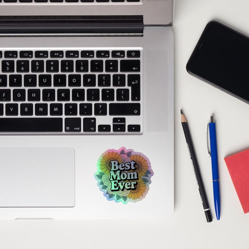 Laptop with 'Best Mom Ever' sticker, phone, and pens on a white surface