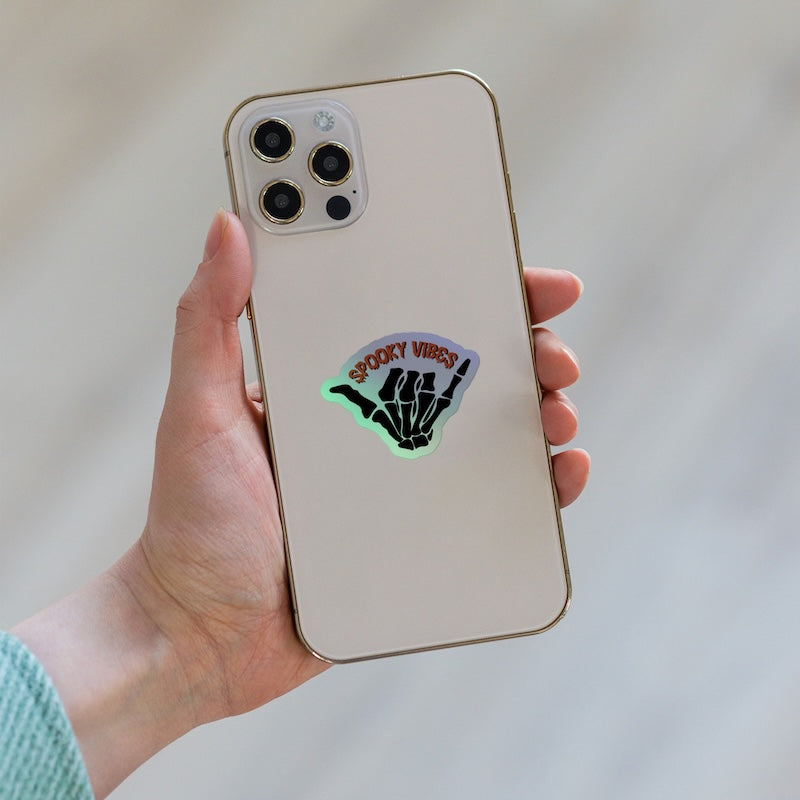 Hand holding a phone with a 'Spooky Vibes' sticker on a light background