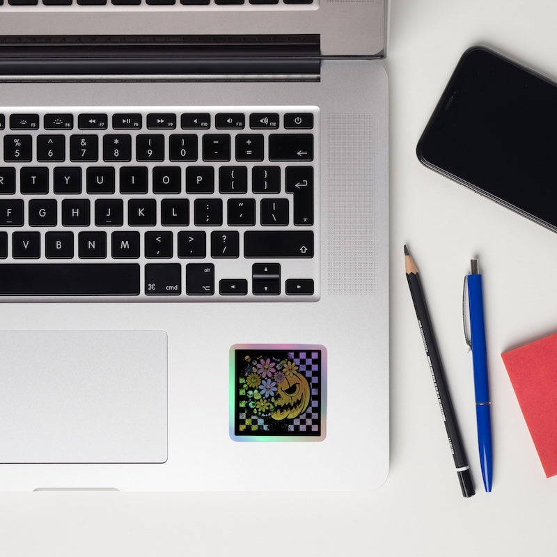 Laptop with a colorful sticker on a white surface, next to a smartphone, pencil, pen, and notebook.