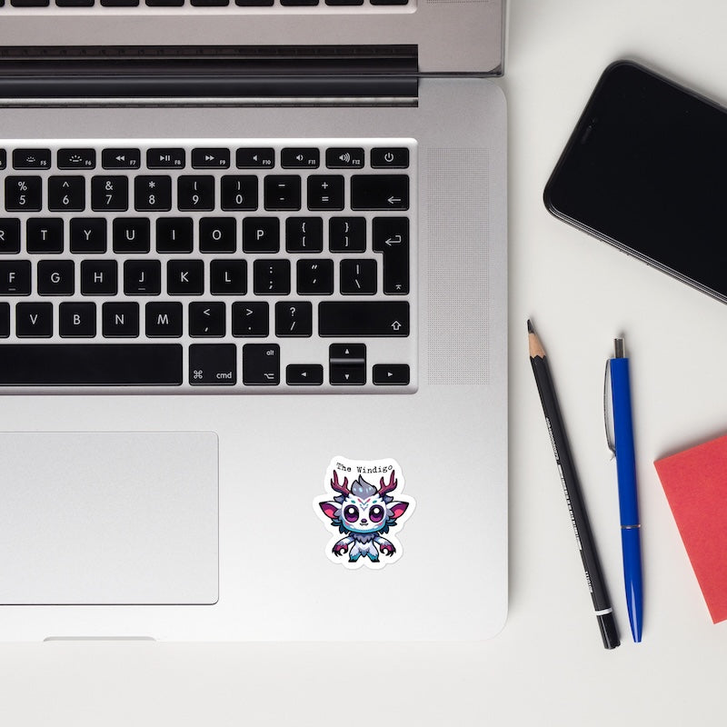 Laptop with a sticker on the palm rest area, next to a smartphone, pencil, pen, and notebook on a white surface.