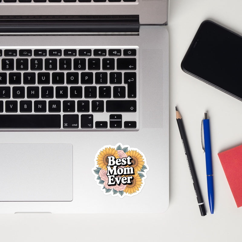 Laptop with 'Best Mom Ever' sticker, phone, pencil, pen, and notebook on a white surface