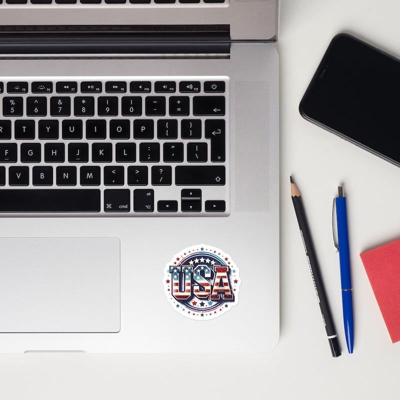 Laptop with a 'USA' sticker, phone, pencil, pen, and notebook on a white surface
