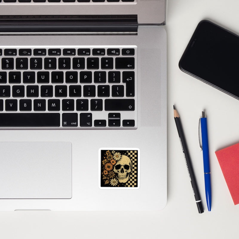 Laptop with a skull and floral design sticker, pencil, pen, and phone on a white surface