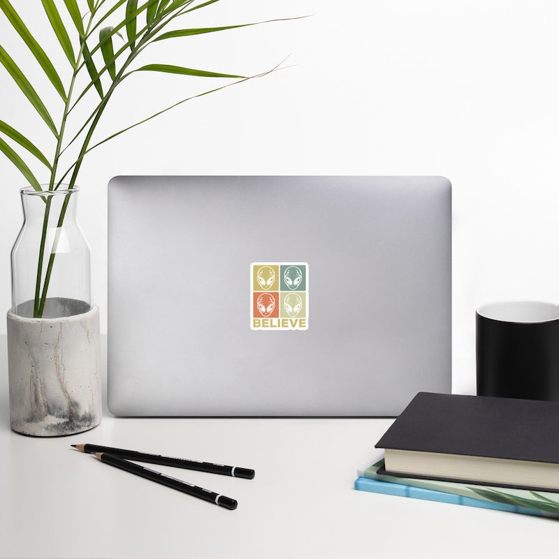 Laptop with a 'BELIEVE' sticker on a desk with a plant, pencils, and books.