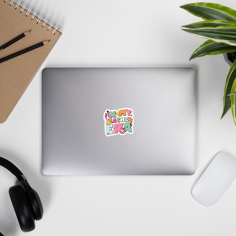 Laptop with a colorful sticker on a desk with a notebook, pencils, headphones, and a plant.