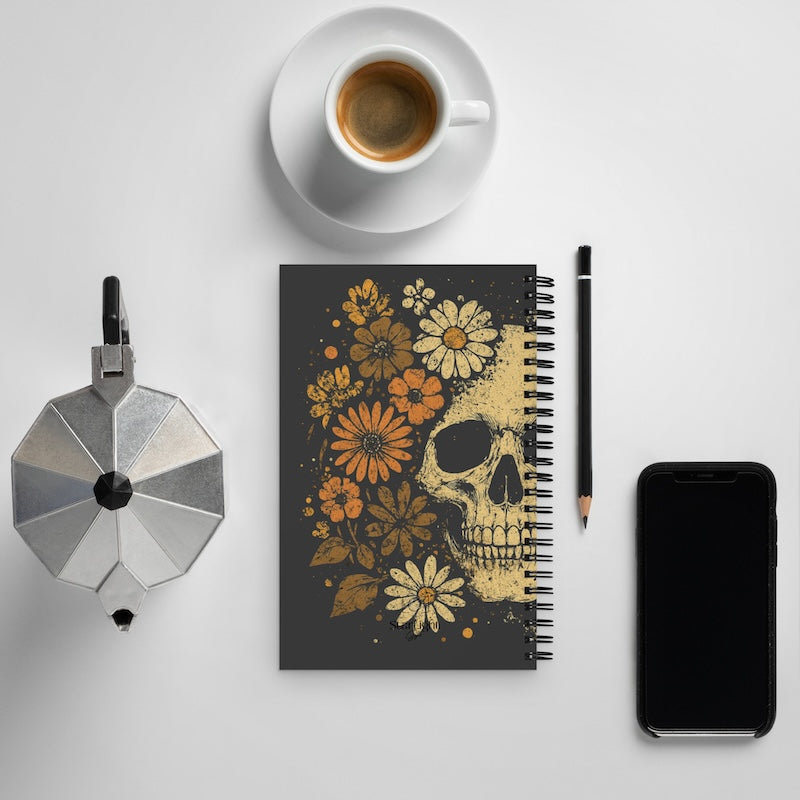 Notebook with skull and floral design on a white surface with coffee, phone, and pencil.