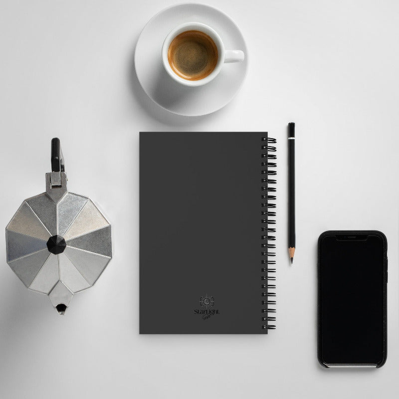 Black notebook with a pen, smartphone, coffee cup, and small metallic object on a light gray background