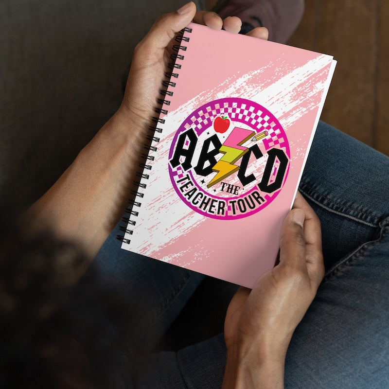 Pink notebook with 'AB CD The Teacher Tour' design held by a person.