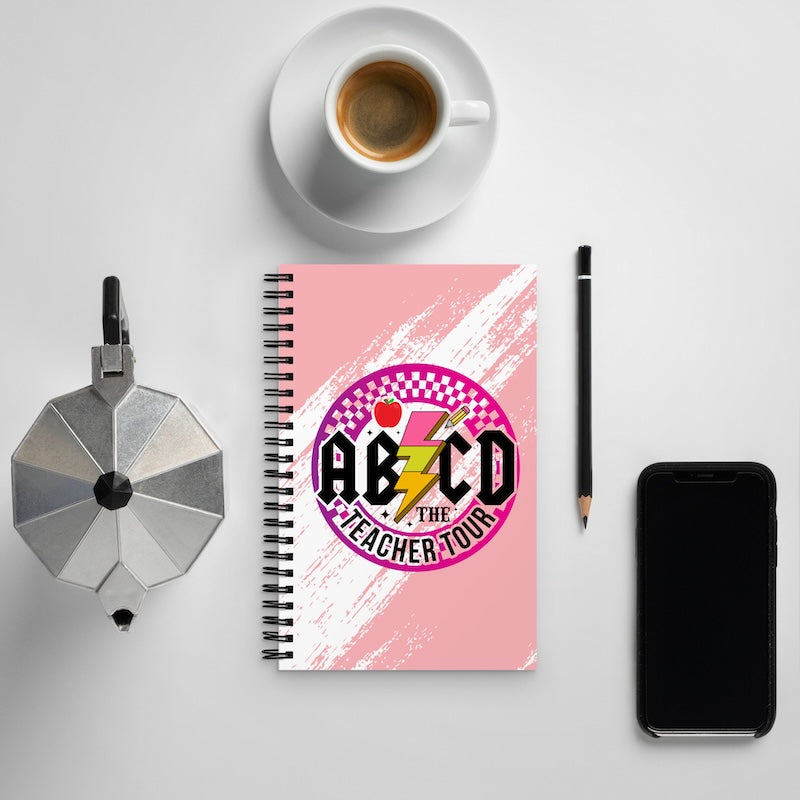Notebook with 'ABZCD The Teacher Tour' design on a white surface with a cup of coffee, phone, and pencil.