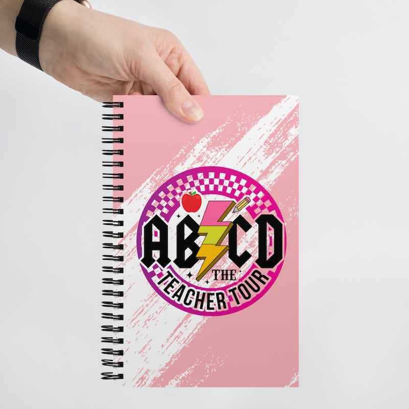 Notebook with 'ABCD The Teacher Tour' design held by a hand on a light gray background