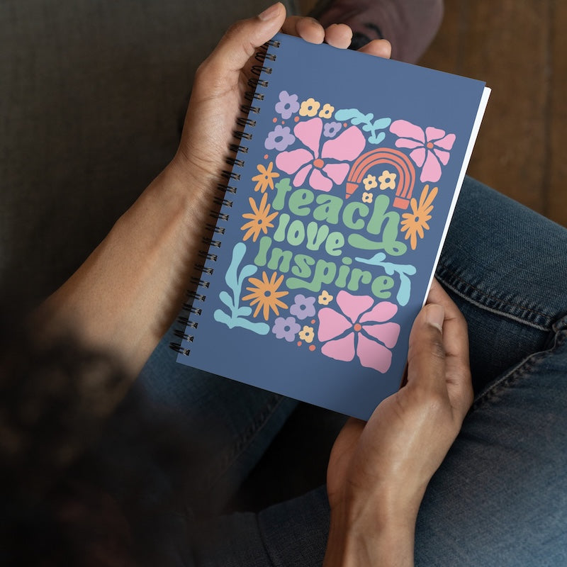 Notebook with 'Teach Love Inspire' design held by hands against a blurred background