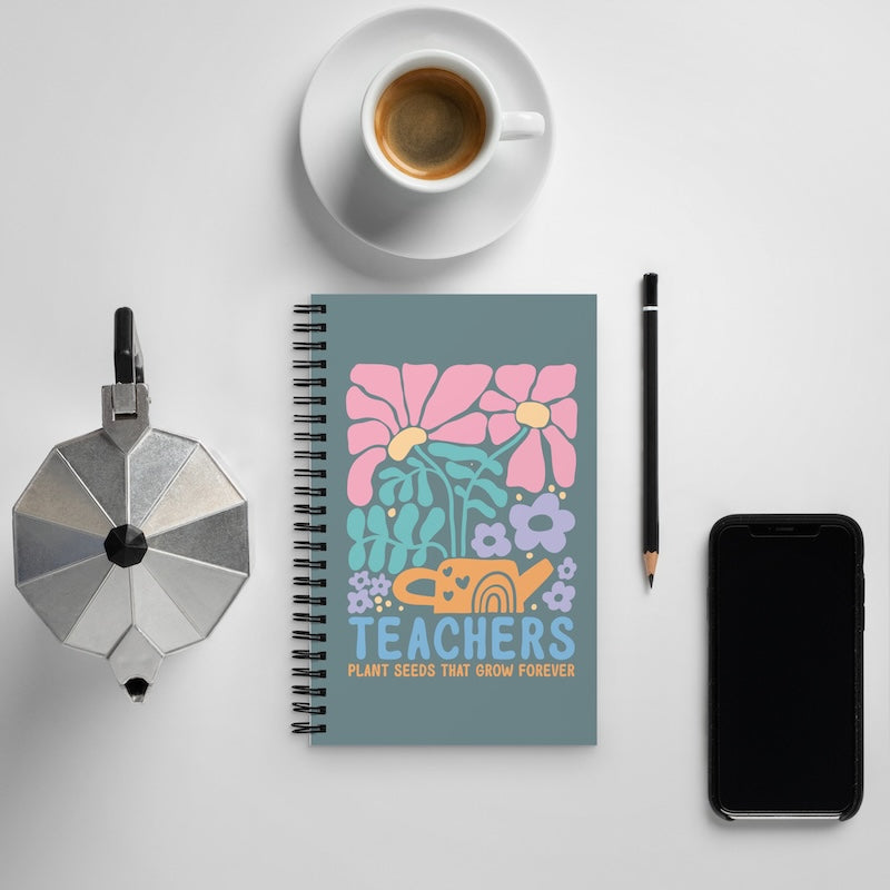Notebook with floral design and 'Teachers Plant Seeds That Grow Forever' text, surrounded by a cup of coffee, phone, and other items on a light gray background.