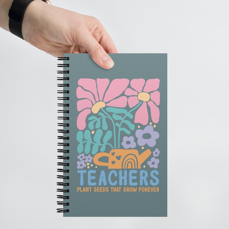 Notebook with floral design and 'Teachers Plant Seeds That Grow Forever' text held by a hand on a light gray background