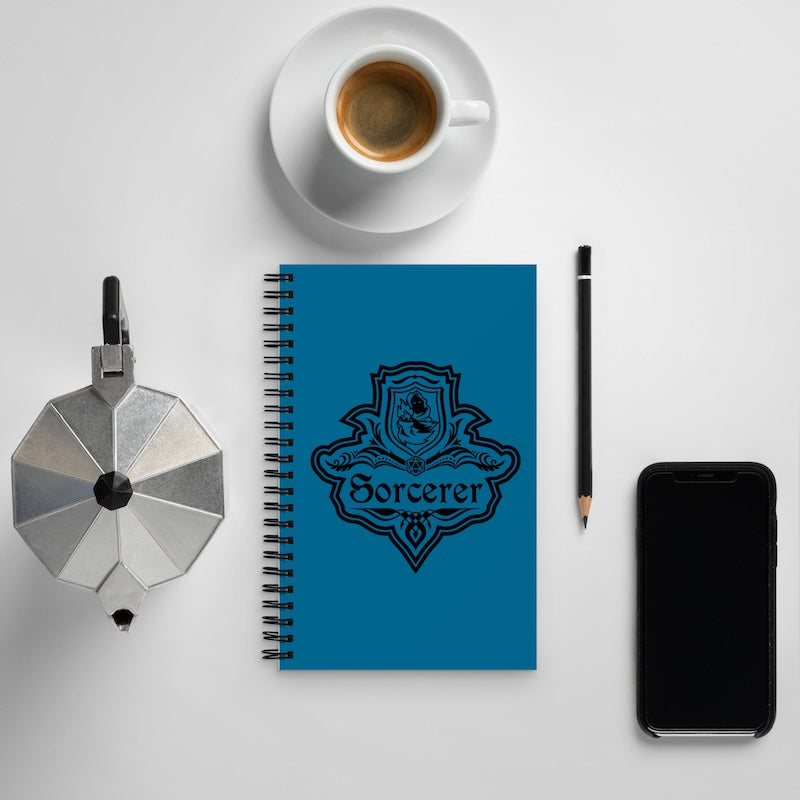 Blue notebook with 'Sorcerer' design, coffee cup, pen, and phone on a white surface