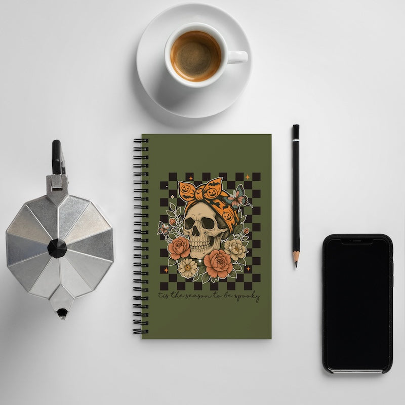 Notebook with skull and floral design on a white surface with a cup, phone, and pen.