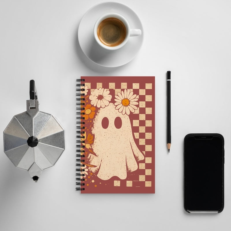Notebook with ghost and flower design on a table with coffee, phone, and other items.