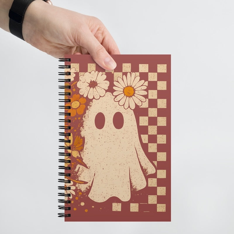Notebook with a ghost and floral design held by a hand on a light gray background