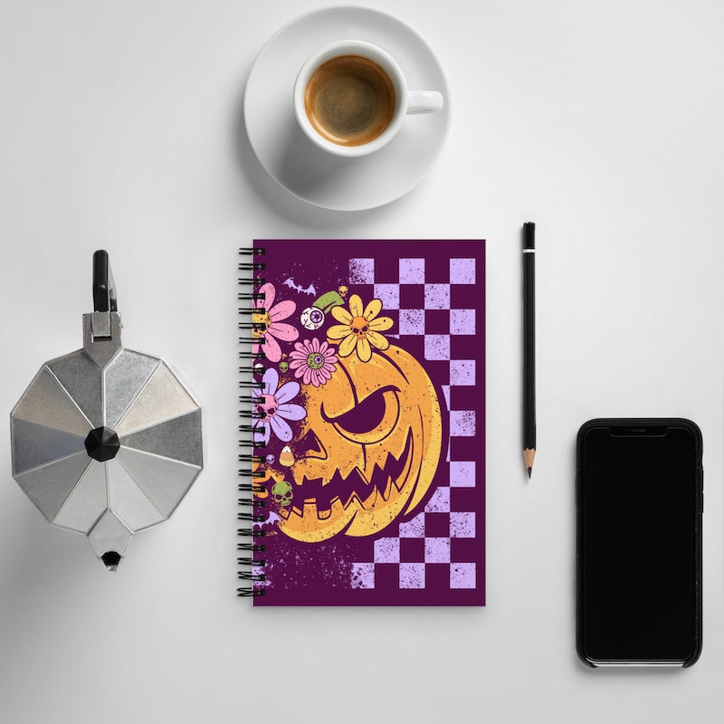 Notebook with pumpkin and floral design on a checkered background, surrounded by a cup of coffee, phone, and other items.