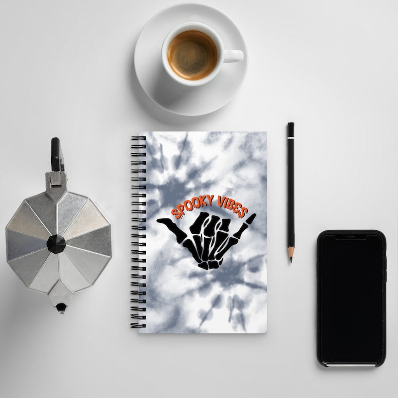 Notebook with 'Spooky Vibes' design, coffee cup, phone, and pen on a light gray background
