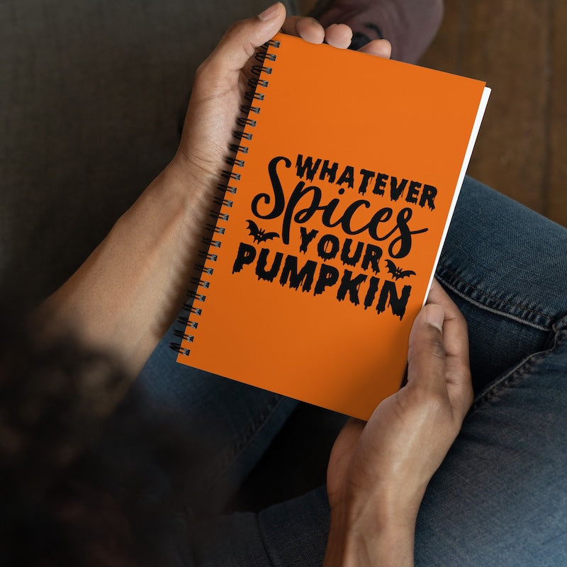 Orange notebook with 'Whatever Spices Your Pumpkin' text held by hands against a dark background