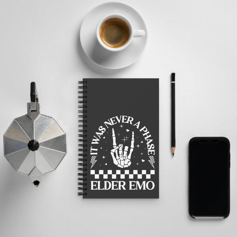 Notebook with 'It was never a phase, Elder Emo' design on a white surface with coffee and phone.