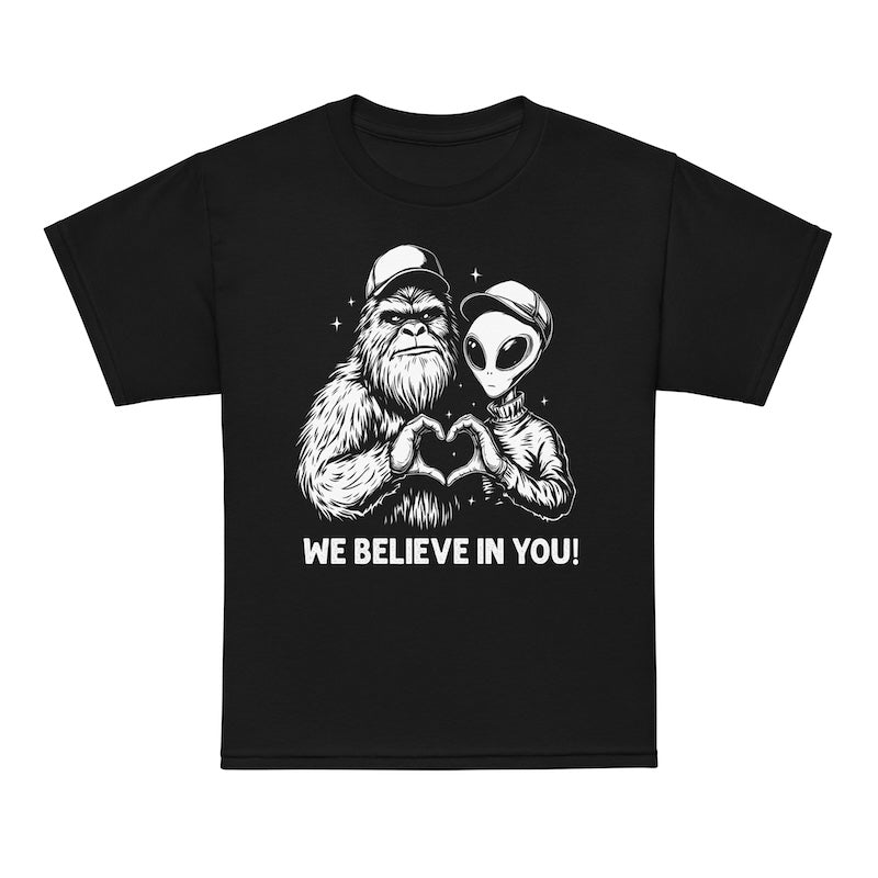 Black t-shirt with a graphic of a bigfoot and alien holding hands and 'WE BELIEVE IN YOU!' text on a white background.
