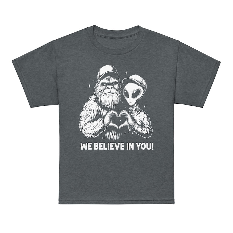 Gray t-shirt with a graphic of a bigfoot and alien holding hands and the text 'We Believe In You!' on a white background.