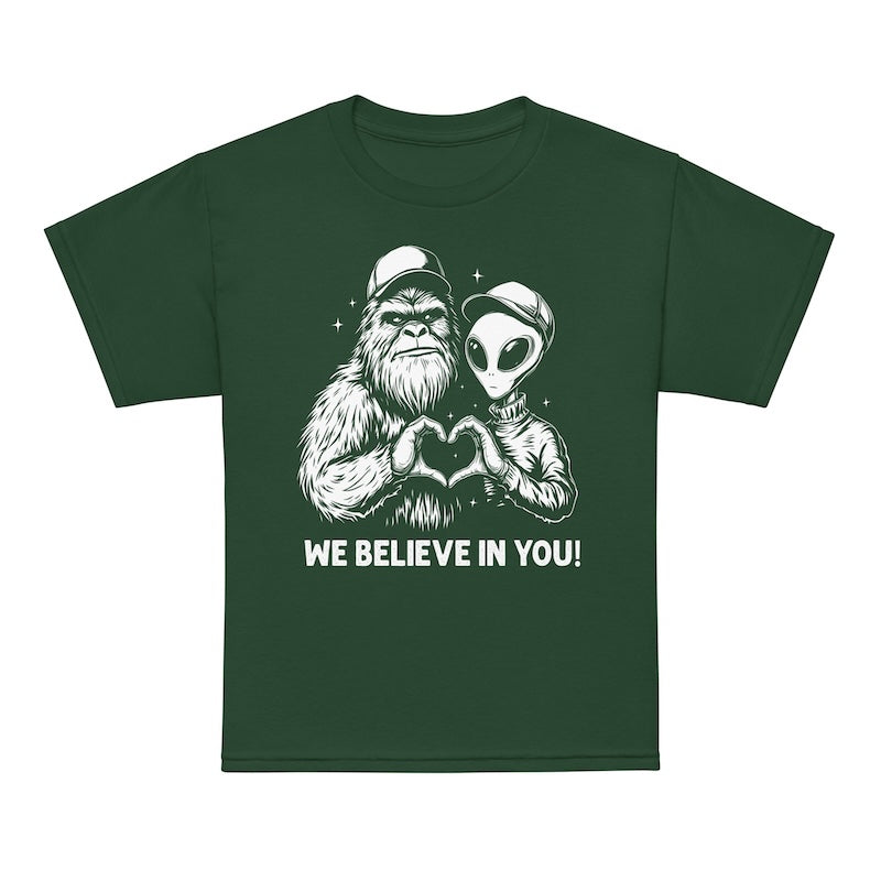 Green t-shirt with a graphic of a bigfoot and alien holding hands and text 'WE BELIEVE IN YOU!' on a white background.